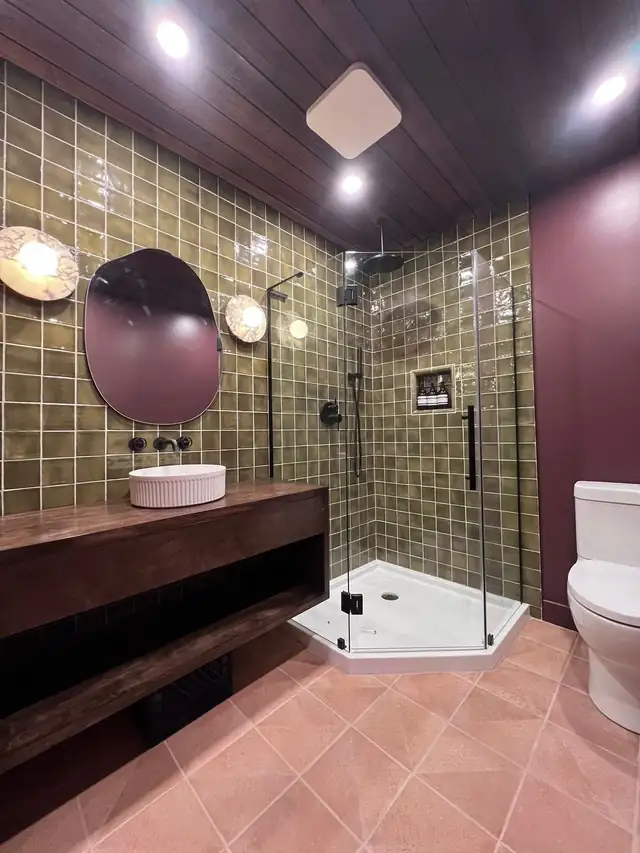 Bathroom remodel with olive green tile, a round mirror, black fixtures, a wood vanity and a dark wood plank ceiling