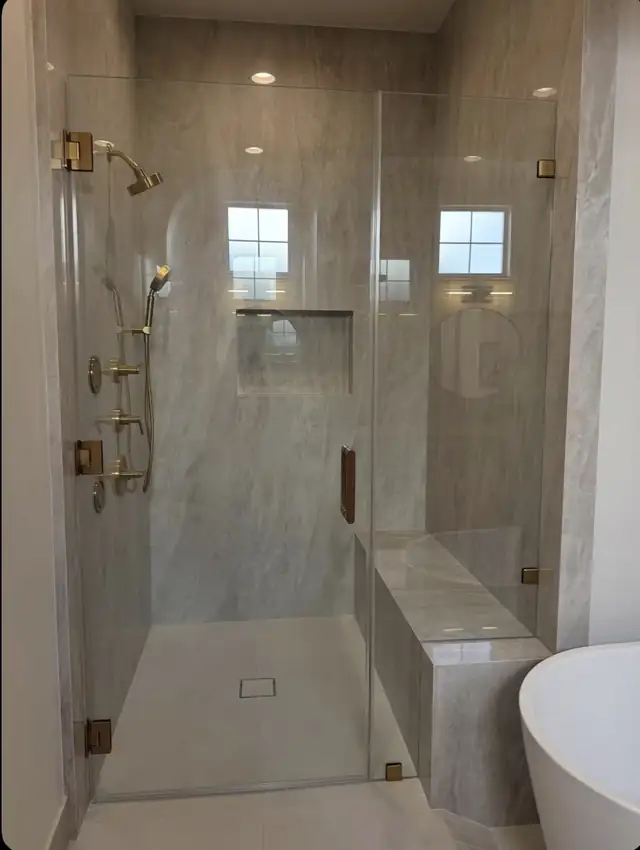 Frameless glass walk-in shower with brass fixtures and tiled bench beside a freestanding tub